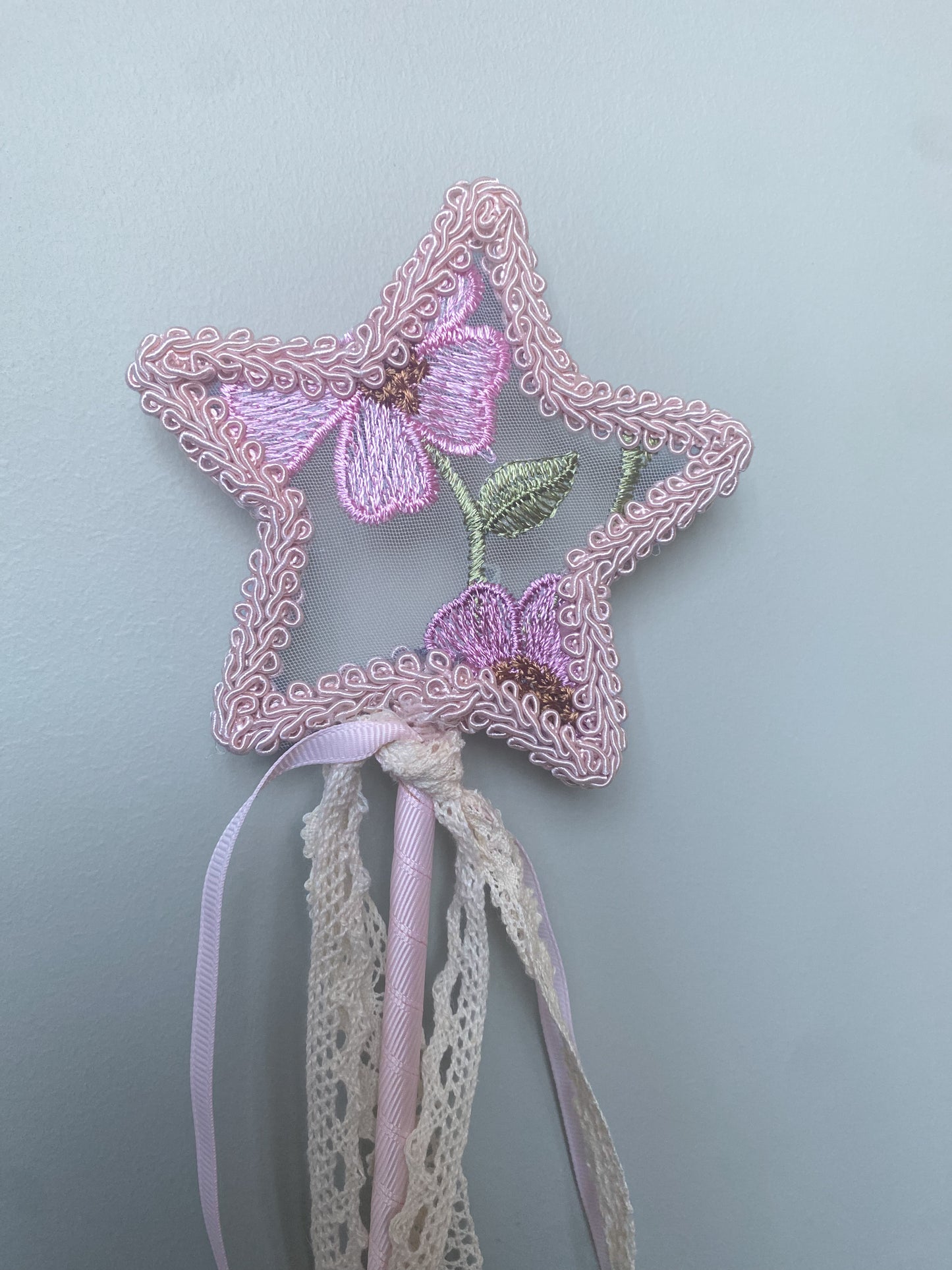 A pink fairy wand with a star shape top embroidered with flowers, attached to a ribbon handle and tassel.
