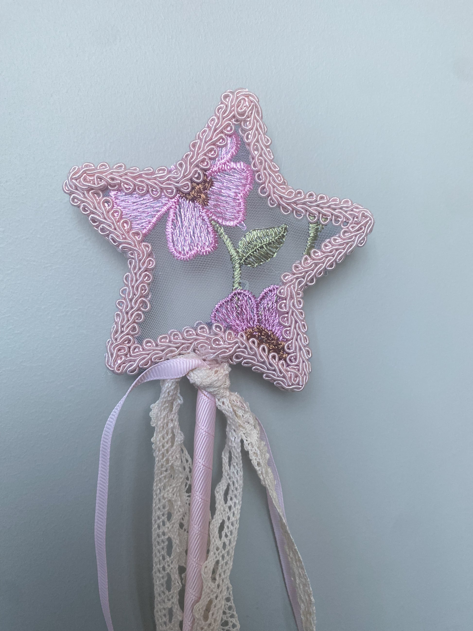A pink fairy wand with a star shape top embroidered with flowers, attached to a ribbon handle and tassel.